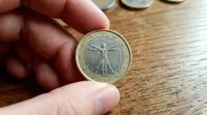 Una mano tiene una moneta da 1 euro con l'Uomo Vitruviano su sfondo di legno